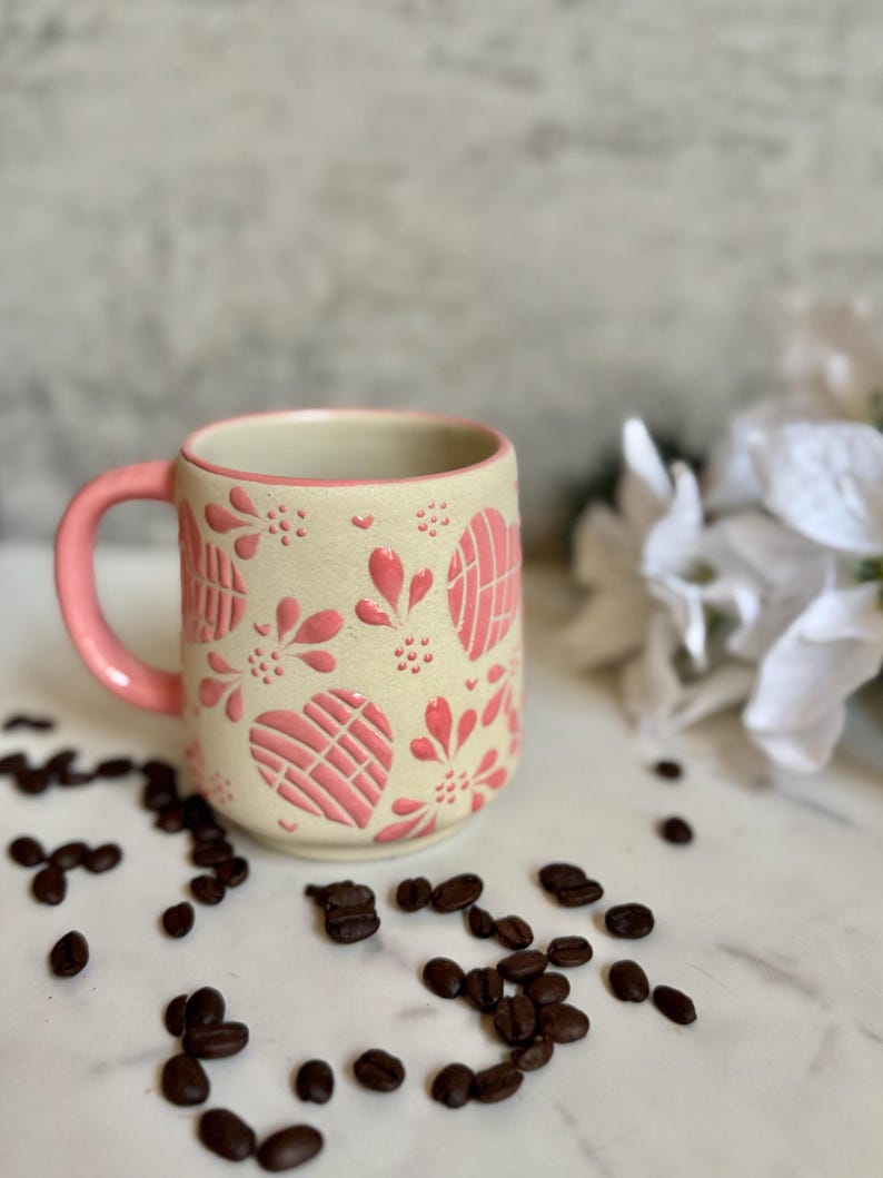 May include: A ceramic coffee mug with a cream-colored base and a pink handle. The mug is decorated with pink heart and floral patterns. Coffee beans are scattered around the mug on a white surface.