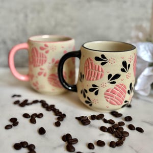 May include: Two ceramic mugs with a cream-colored base, adorned with pink heart-shaped designs and black floral accents. One mug has a pink handle, the other a black handle. Coffee beans are scattered around the mugs.