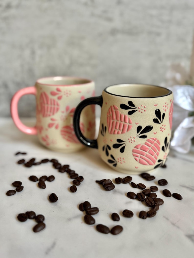 May include: Two ceramic mugs with a cream-colored base, adorned with pink heart-shaped designs and black floral accents. One mug has a pink handle, the other a black handle. Coffee beans are scattered around the mugs.