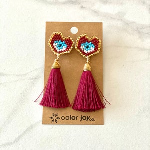 May include: A pair of red heart-shaped earrings with blue and white evil eye beading and burgundy tassel drops. The earrings are displayed on a brown card with the text "color joy co."