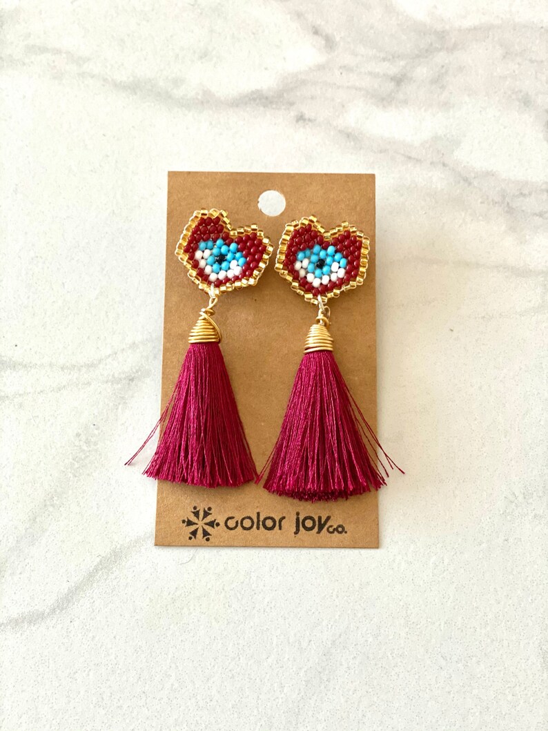 May include: A pair of red heart-shaped earrings with blue and white evil eye beading and burgundy tassel drops. The earrings are displayed on a brown card with the text "color joy co."