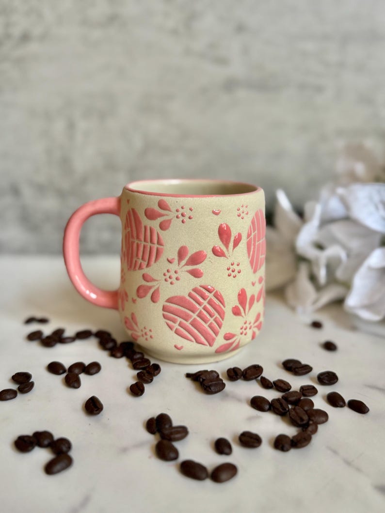 May include: A ceramic coffee mug with a cream-colored base and a pink handle. The mug is decorated with pink floral and heart-shaped patterns. Coffee beans are scattered around the mug, suggesting a coffee-related theme.