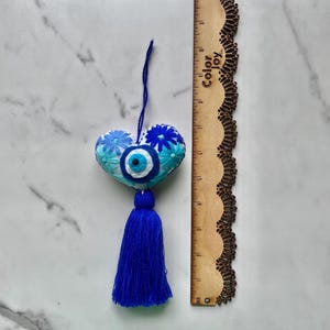 May include: White heart-shaped ornament with blue and turquoise embroidered flowers and a blue evil eye charm. The ornament has a blue tassel.
