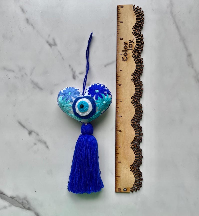 May include: White heart-shaped ornament with blue and turquoise embroidered flowers and a blue evil eye charm. The ornament has a blue tassel.