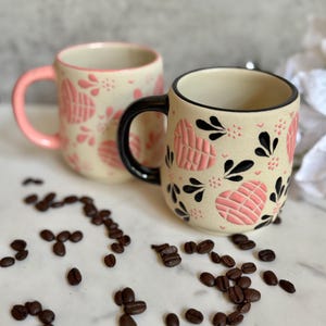 May include: Two ceramic mugs with a cream-colored base, adorned with pink heart-shaped patterns and black accents. One mug has a pink handle, the other a black handle. Coffee beans are scattered around the mugs.