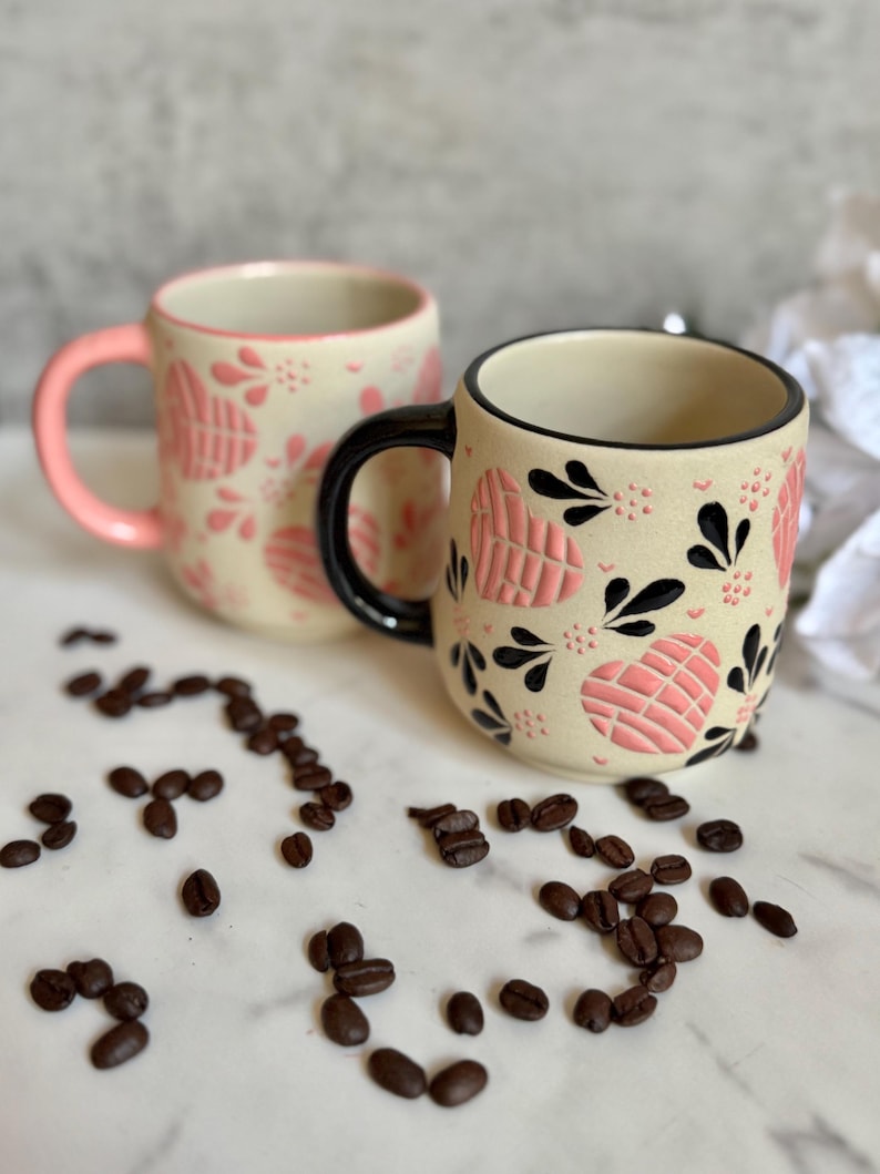 May include: Two ceramic mugs with a cream-colored base, adorned with pink heart-shaped patterns and black accents. One mug has a pink handle, the other a black handle. Coffee beans are scattered around the mugs.