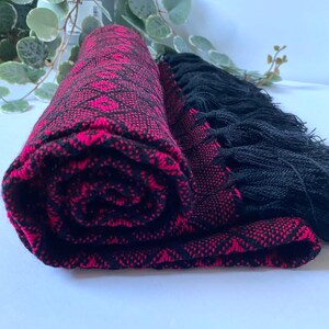 May include: A pink and black woven shawl with black fringe. The shawl is rolled up and the fringe is visible.