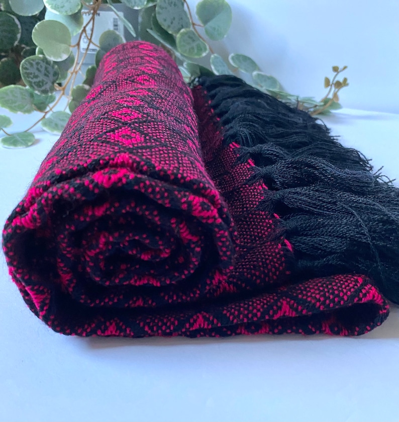May include: A pink and black woven shawl with black fringe. The shawl is rolled up and the fringe is visible.