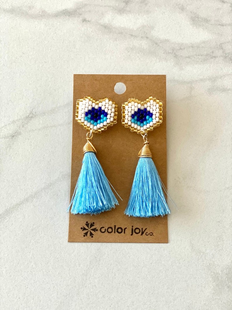 May include: A pair of blue tassel earrings with gold accents. The earrings feature a white and blue beaded evil eye design.