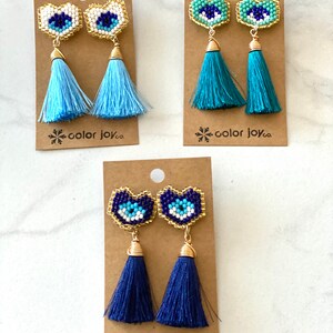 May include: Three pairs of heart-shaped earrings with blue evil eye charms and tassel drops. The tassels are light blue, teal, and navy blue. The earrings are displayed on brown cards with the text "color joy co."