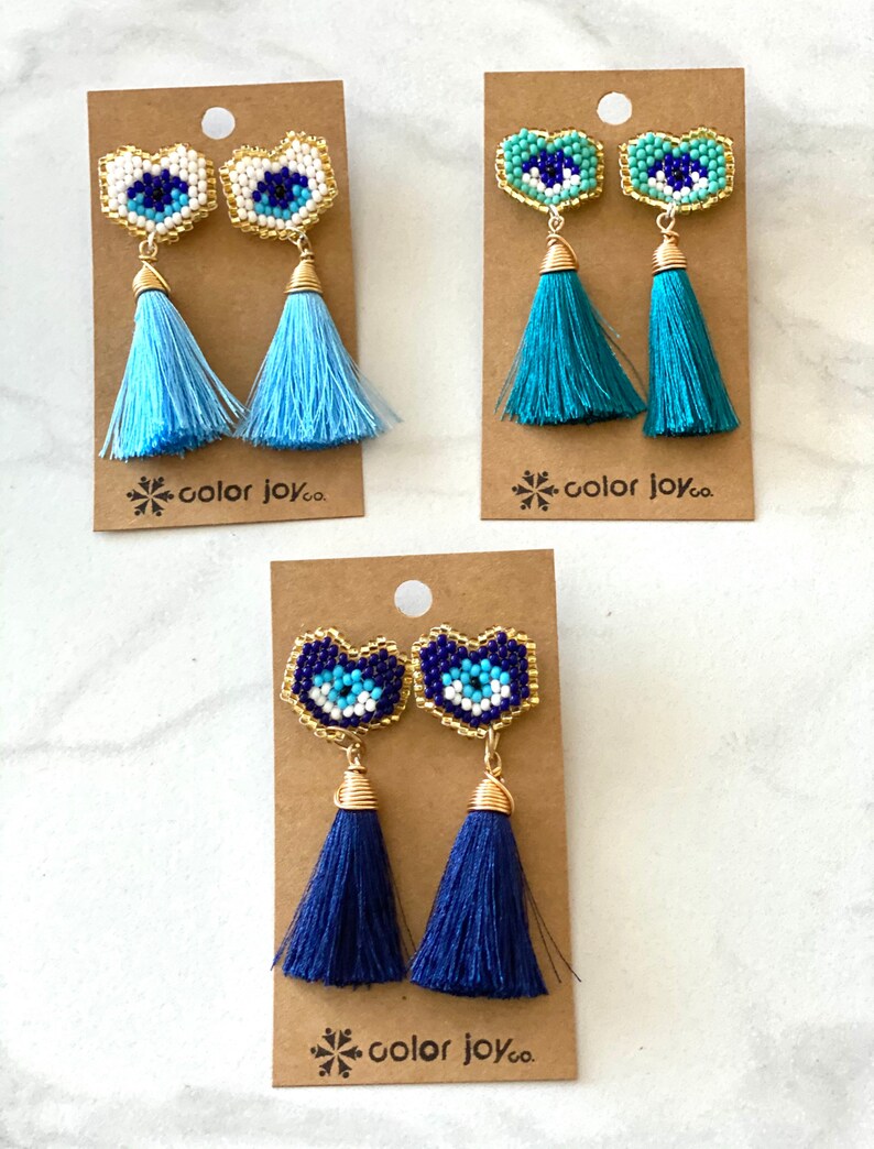 May include: Three pairs of heart-shaped earrings with blue evil eye charms and tassel drops. The tassels are light blue, teal, and navy blue. The earrings are displayed on brown cards with the text "color joy co."