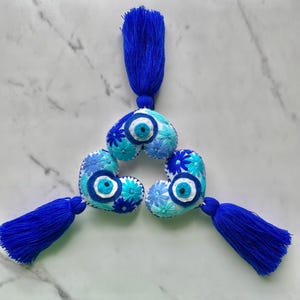 May include: Three blue and white heart-shaped embroidered ornaments with blue tassels. Each heart features a blue evil eye charm and blue and white floral embroidery.
