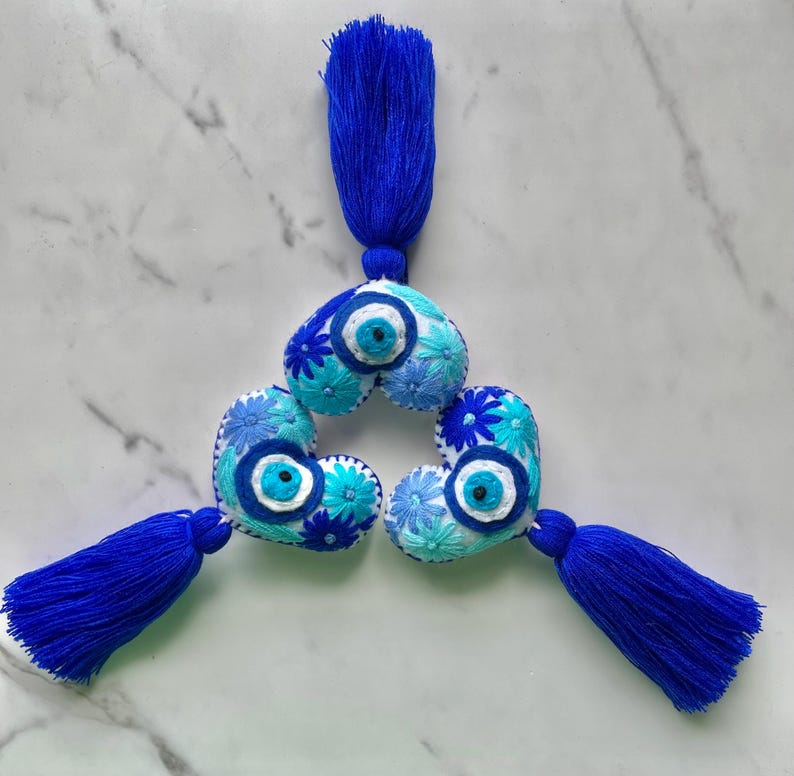 May include: Three blue and white heart-shaped embroidered ornaments with blue tassels. Each heart features a blue evil eye charm and blue and white floral embroidery.