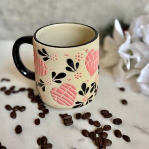 May include: A ceramic coffee mug with a cream-colored base, adorned with pink heart-shaped designs and black floral accents. The mug has a black handle and rim. Coffee beans are scattered around the mug on a white surface.