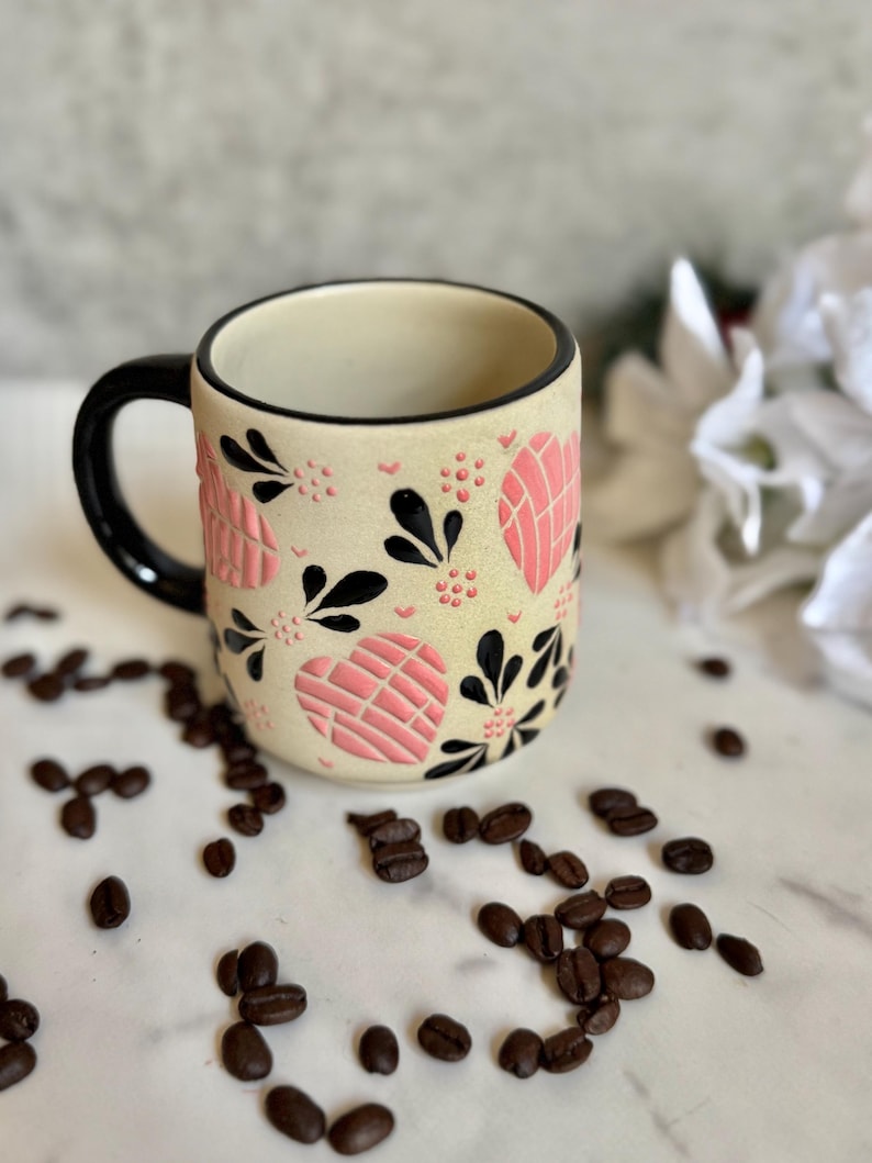 May include: A ceramic coffee mug with a cream-colored base, adorned with pink heart-shaped designs and black floral accents. The mug has a black handle and rim. Coffee beans are scattered around the mug on a white surface.