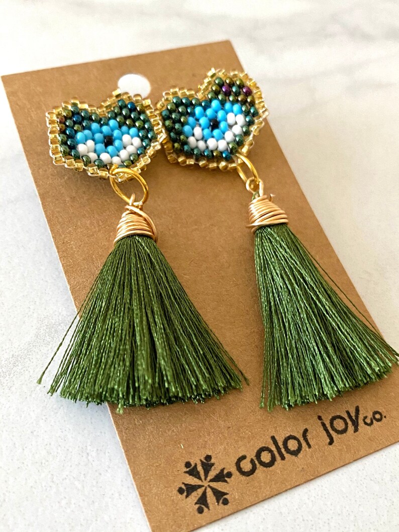 May include: A pair of green tassel earrings with a beaded heart-shaped charm featuring a blue evil eye design. The earrings are on a brown card with the text "color joy co."