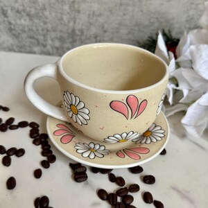 May include: A beige ceramic teacup and saucer set decorated with white daisies and pink accents. The teacup has a curved handle and sits on a matching saucer. Coffee beans are scattered around the set.