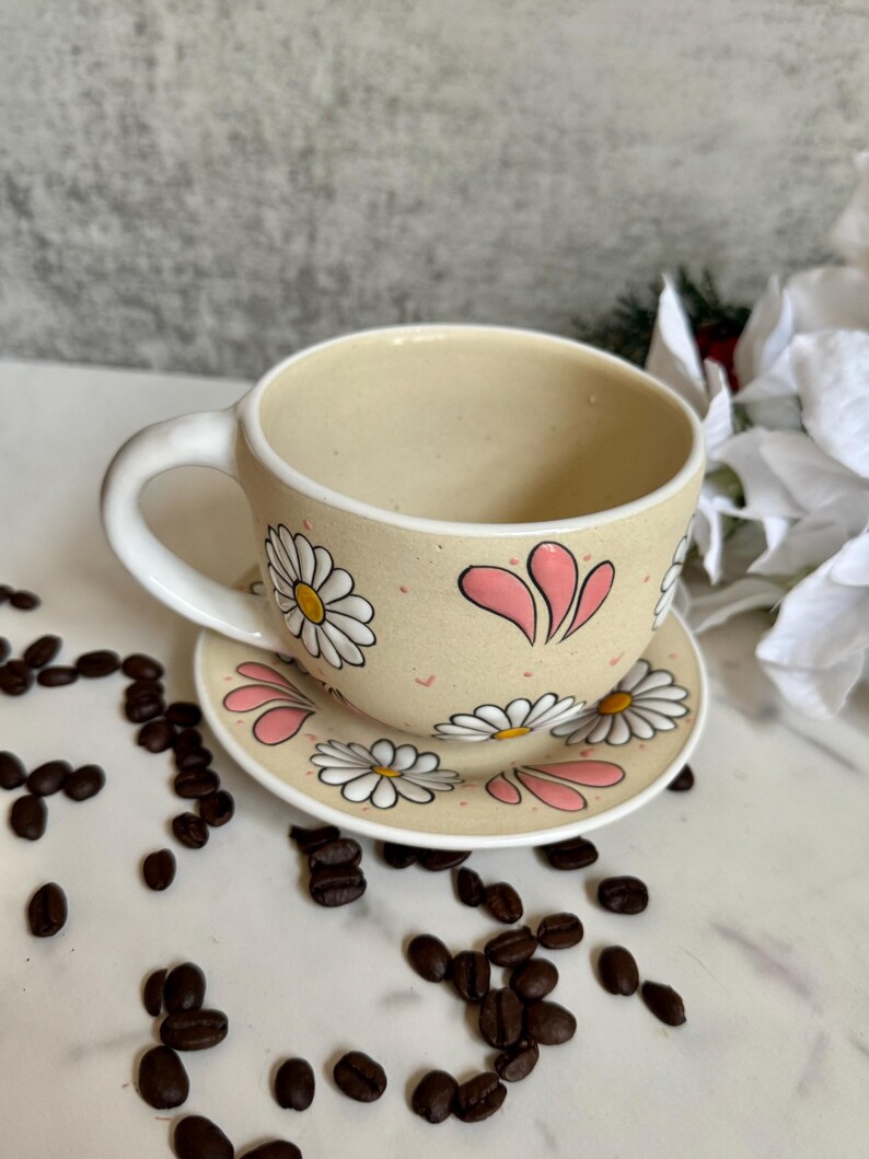 May include: A beige ceramic teacup and saucer set decorated with white daisies and pink accents. The teacup has a curved handle and sits on a matching saucer. Coffee beans are scattered around the set.