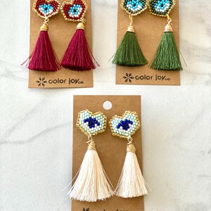 May include: Three pairs of beaded heart-shaped earrings with a blue evil eye design. Each pair has a different colored tassel: burgundy, green, and cream.
