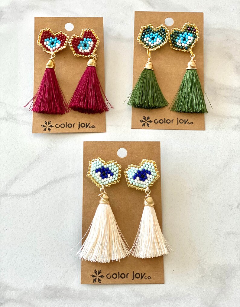May include: Three pairs of beaded heart-shaped earrings with a blue evil eye design. Each pair has a different colored tassel: burgundy, green, and cream.