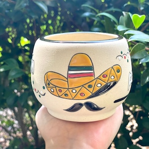 May include: A white ceramic planter with a black rim and a hand-painted design of a yellow sombrero with green, red, and white stripes, a black mustache, and white dots.