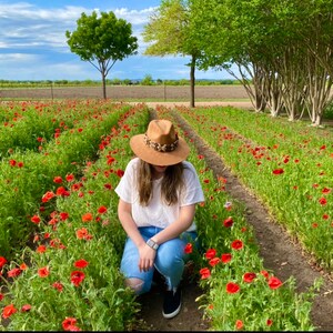 May include: A person wearing a brown hat sits in a field of red poppies. The field is surrounded by green trees and a blue sky.