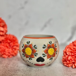 May include: A ceramic sugar skull-themed bowl with orange and yellow floral designs. The bowl features a black heart-shaped nose, red lips, and blue dots. The bowl is surrounded by orange flowers.