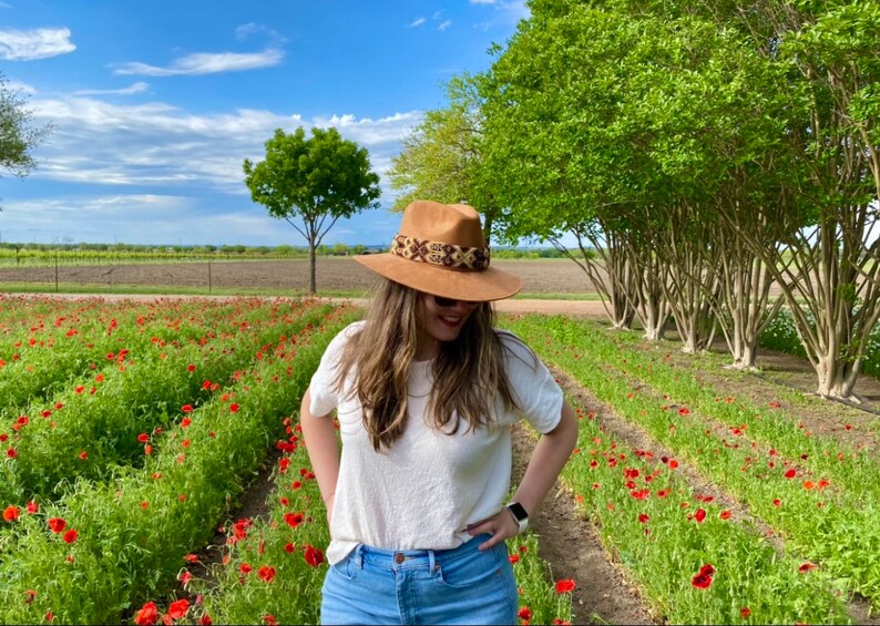 May include: A woman wearing a white t-shirt and blue jeans stands in a field of red poppies. She is wearing a brown hat with a patterned band.
