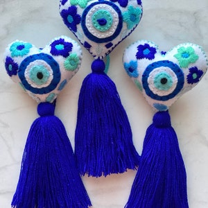 May include: Three white felt heart-shaped ornaments with blue and green embroidered flowers and a blue evil eye charm. Each ornament has a blue tassel.