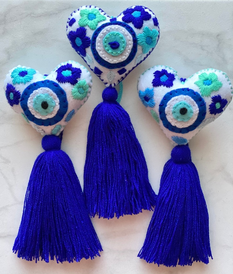 May include: Three white felt heart-shaped ornaments with blue and green embroidered flowers and a blue evil eye charm. Each ornament has a blue tassel.