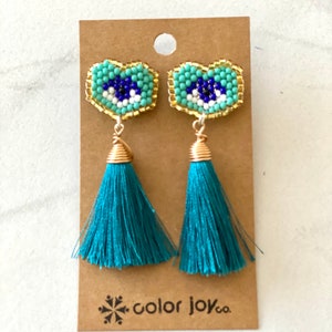May include: Teal tassel earrings with a blue and white evil eye charm. The charms are surrounded by gold beads and the earrings are on a brown card with the text "color joy co."
