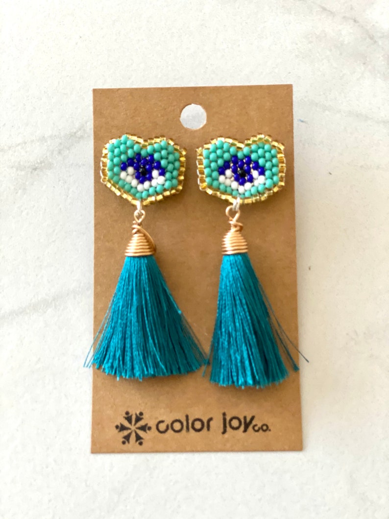 May include: Teal tassel earrings with a blue and white evil eye charm. The charms are surrounded by gold beads and the earrings are on a brown card with the text "color joy co."