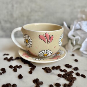 May include: A beige ceramic teacup and saucer set decorated with white daisies and pink accents. The cup has a curved handle and sits on a matching saucer. Coffee beans are scattered around the set.