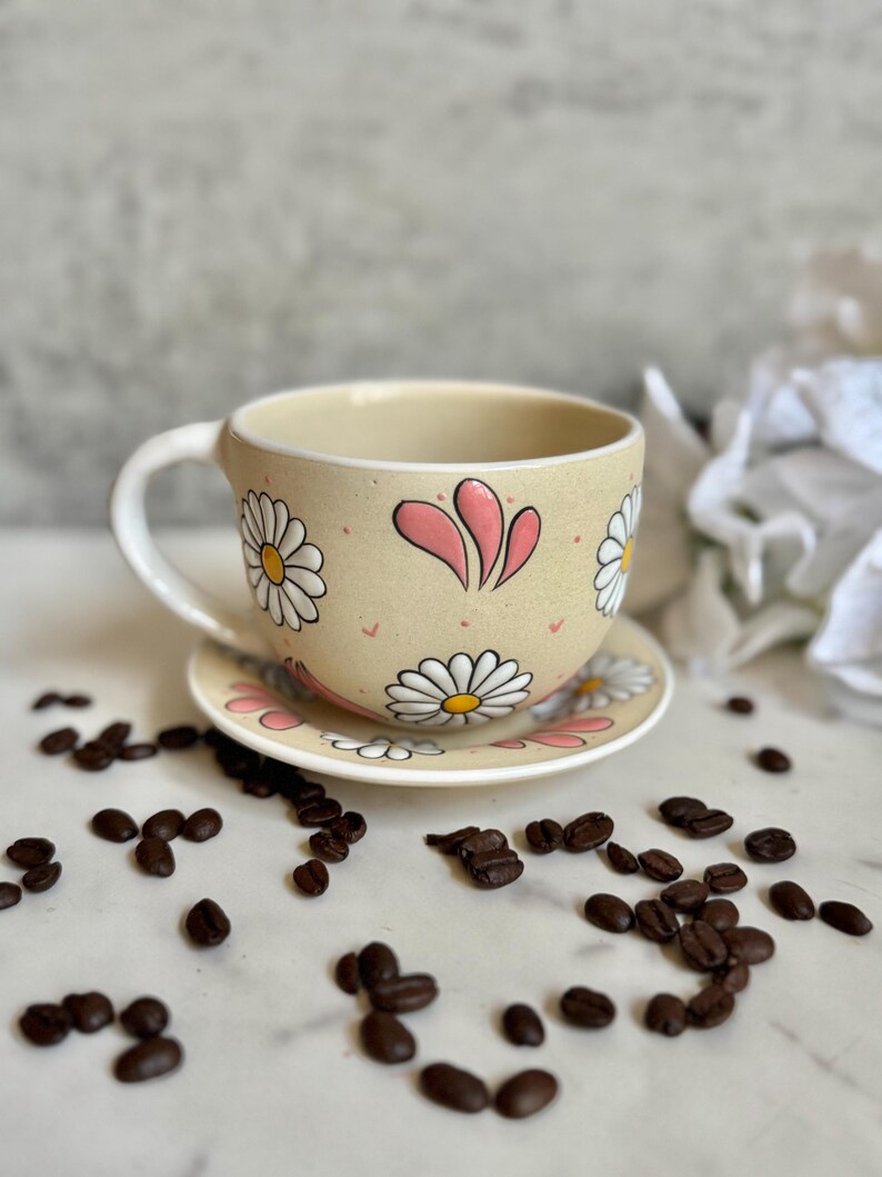 May include: A beige ceramic teacup and saucer set decorated with white daisies and pink accents. The cup has a curved handle and sits on a matching saucer. Coffee beans are scattered around the set.