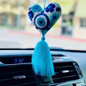 May include: Blue and white felt heart with an evil eye charm and a blue tassel hanging from the rearview mirror of a car. The car's digital clock reads 7:32.