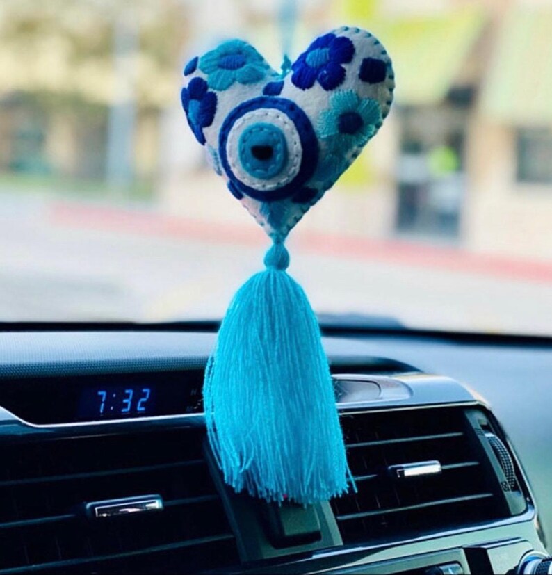 May include: Blue and white felt heart with an evil eye charm and a blue tassel hanging from the rearview mirror of a car. The car's digital clock reads 7:32.