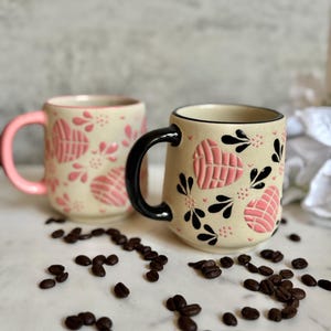 May include: Two ceramic mugs with pink and black accents. The mugs have a beige background with pink heart and floral designs. One mug has a pink handle, and the other has a black handle. Coffee beans are scattered around the mugs.