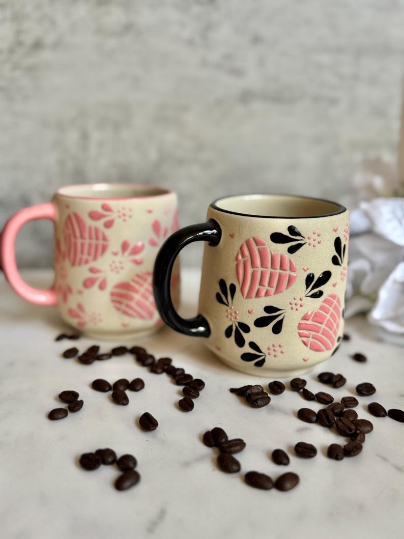 May include: Two ceramic mugs with pink and black accents. The mugs have a beige background with pink heart and floral designs. One mug has a pink handle, and the other has a black handle. Coffee beans are scattered around the mugs.