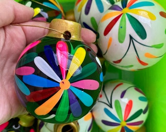 May include: Hand-painted Christmas ornaments with a floral design. The ornaments are round, with a dark green or white base and colorful petals in shades of pink, yellow, blue, and orange. Each ornament has a gold-colored cap and a gold string for hanging.