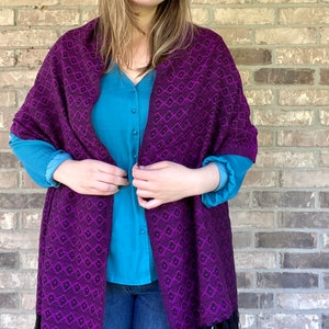May include: A purple and black shawl with a geometric pattern. The shawl is made of a soft, woven fabric and has a fringe border.