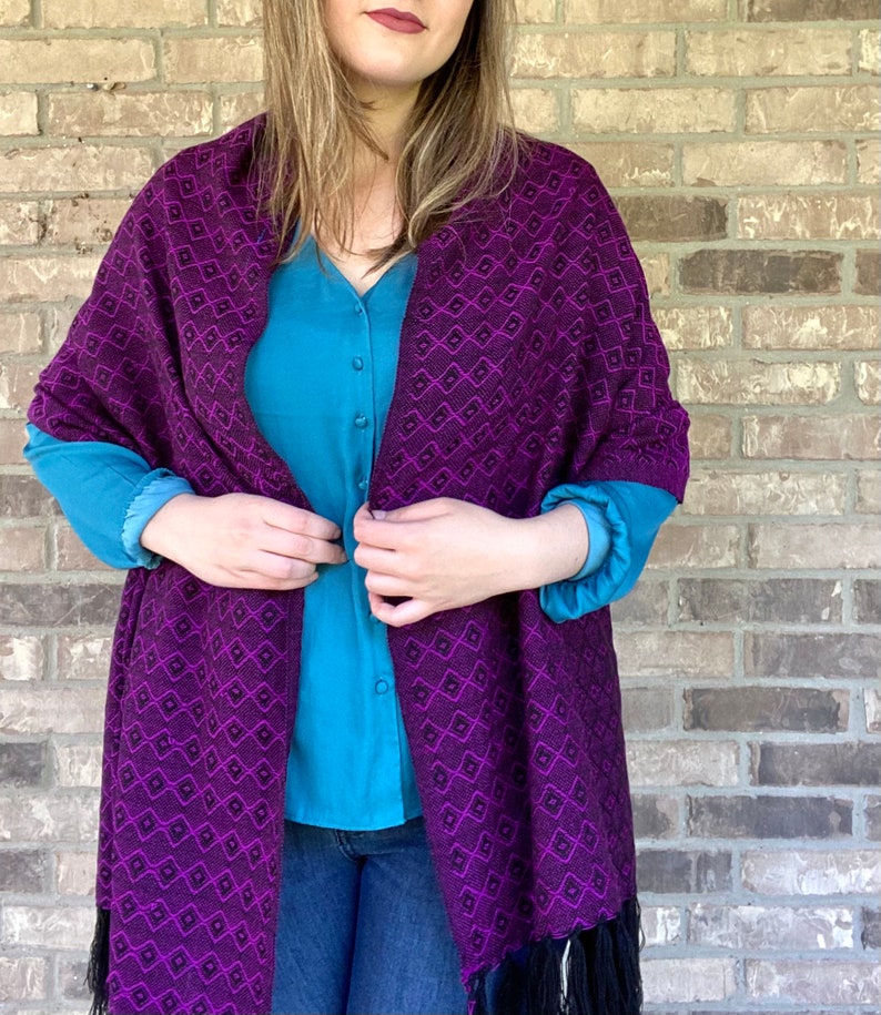 May include: A purple and black shawl with a geometric pattern. The shawl is made of a soft, woven fabric and has a fringe border.