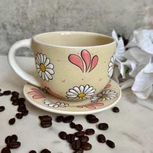 May include: A beige ceramic teacup and saucer set decorated with white daisies and pink accents. The cup has a white handle and is filled with a dark liquid. Coffee beans are scattered around the cup and saucer on a white surface.