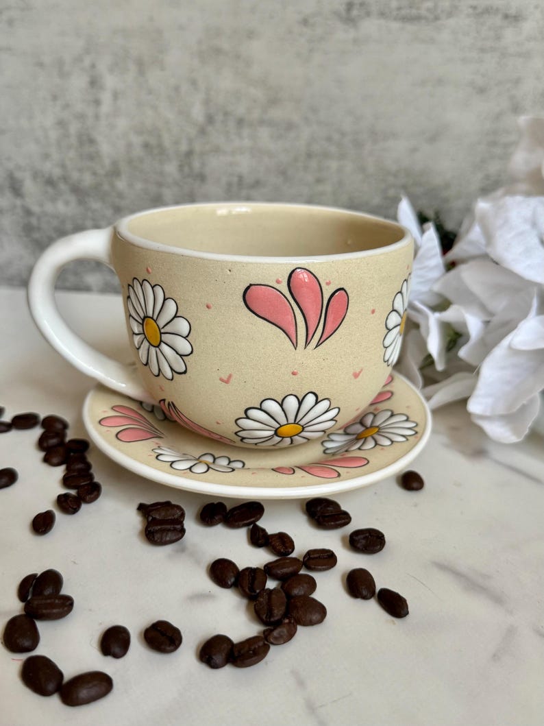 May include: A beige ceramic teacup and saucer set decorated with white daisies and pink accents. The cup has a white handle and is filled with a dark liquid. Coffee beans are scattered around the cup and saucer on a white surface.