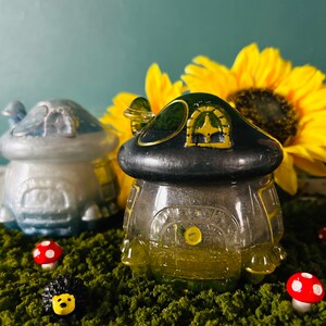 May include: Two miniature houses made of glass and ceramic, one blue and one green, with a yellow sunflower in the background. The houses are sitting on a bed of green moss with red and white mushrooms.