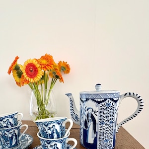 May include: A blue and white porcelain tea set with a teapot, four teacups, and saucers. The set features a detailed illustration of figures and patterns. A vase of orange flowers sits in the background.