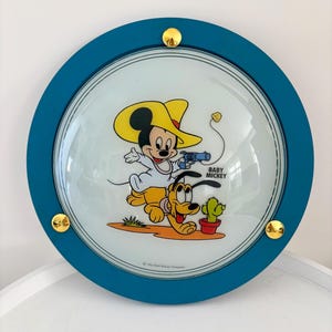 Pode incluir: Uma luminária de parede redonda azul turquesa com um domo de vidro que apresenta uma imagem de desenho animado do Baby Mickey Mouse e Pluto. A imagem mostra o Baby Mickey usando um chapéu de cowboy amarelo e segurando uma arma de brinquedo, enquanto o Pluto corre atrás dele. O texto "BABY MICKEY" está impresso no domo de vidro.