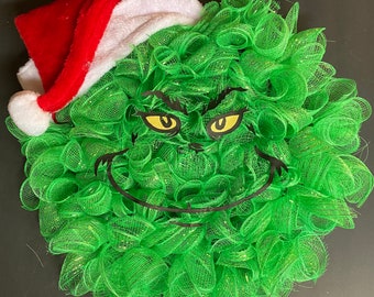 May include: A green wreath with a Grinch face and a red and white Santa hat. The Grinch's eyes are yellow and his mouth is black.