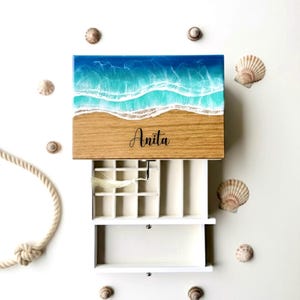 Può includere: Un portagioie in legno con un design a scena di spiaggia. La parte superiore presenta un oceano blu e turchese con onde bianche su una spiaggia beige. Il nome "Anita" è scritto in corsivo. La scatola è aperta, rivelando scomparti bianchi.