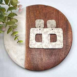 May include: Square-shaped earrings with a neutral, textured pattern. The earrings are a light beige color with a darker beige pattern. They are displayed on a wooden and marble serving board. The earrings have a post and a rectangular cutout.