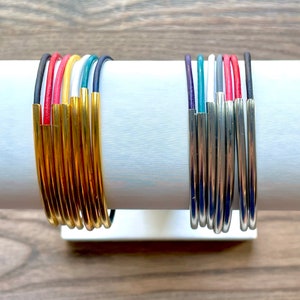 Könnte beinhalten: Ein Satz goldener und silberner Armreifen mit bunten Lederakzenten. Die Armreifen sind auf einem weißen Präsentationsständer gestapelt.